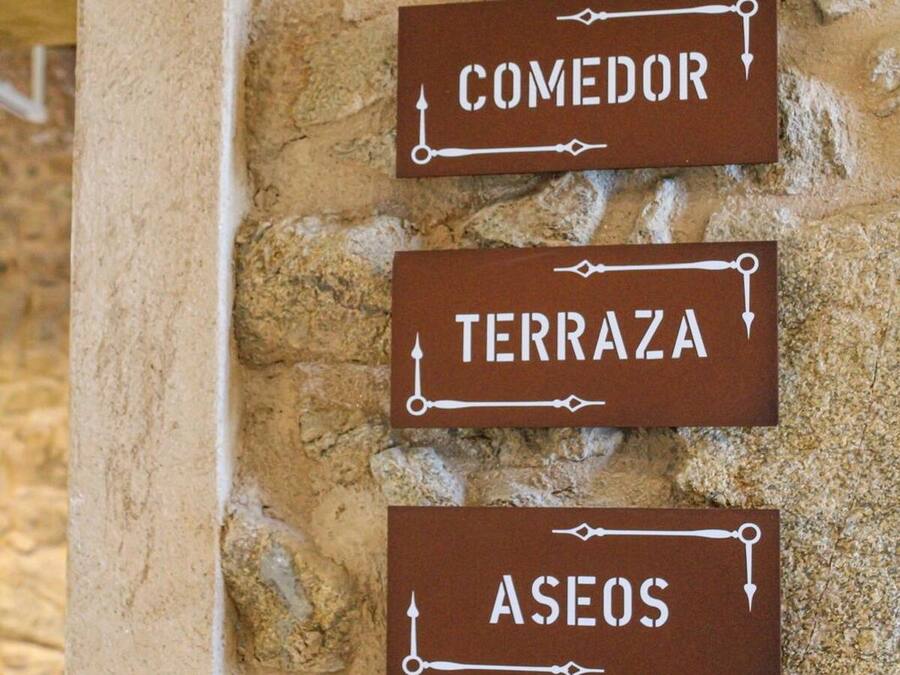 Tres letreros marrones con texto blanco sobre una pared de piedra, con la etiqueta 'COMEDOR', 'TERRAZA' y 'ASEOS'.