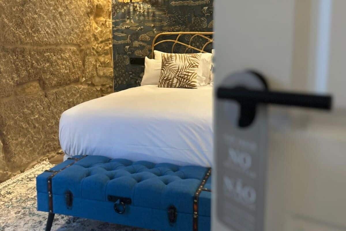 Una cama con ropa blanca, un banco tapizado en azul y una pared de piedra.