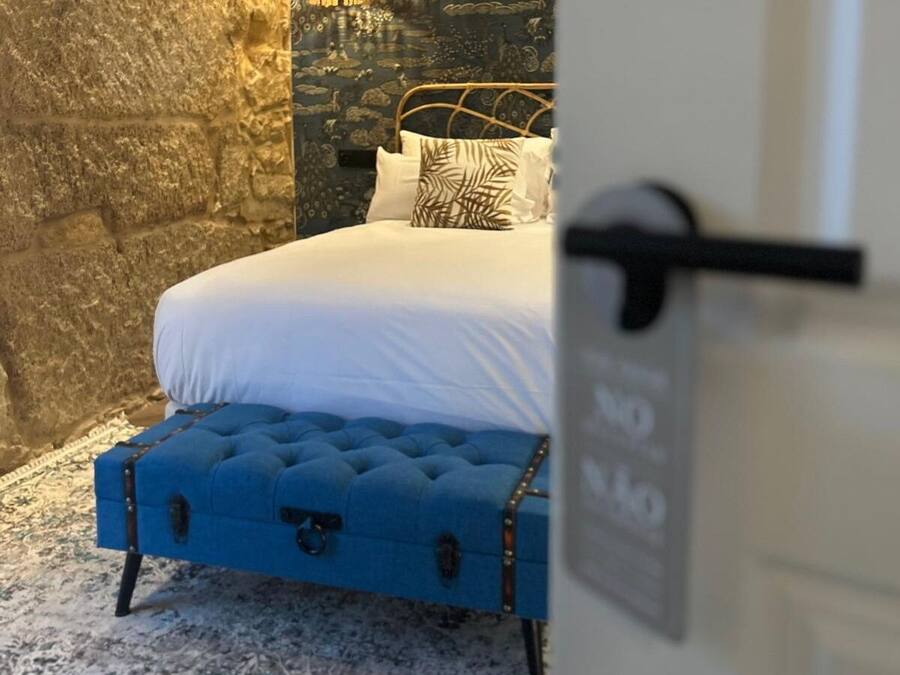 A bed with white bedding, a blue upholstered bench, and a stone wall.