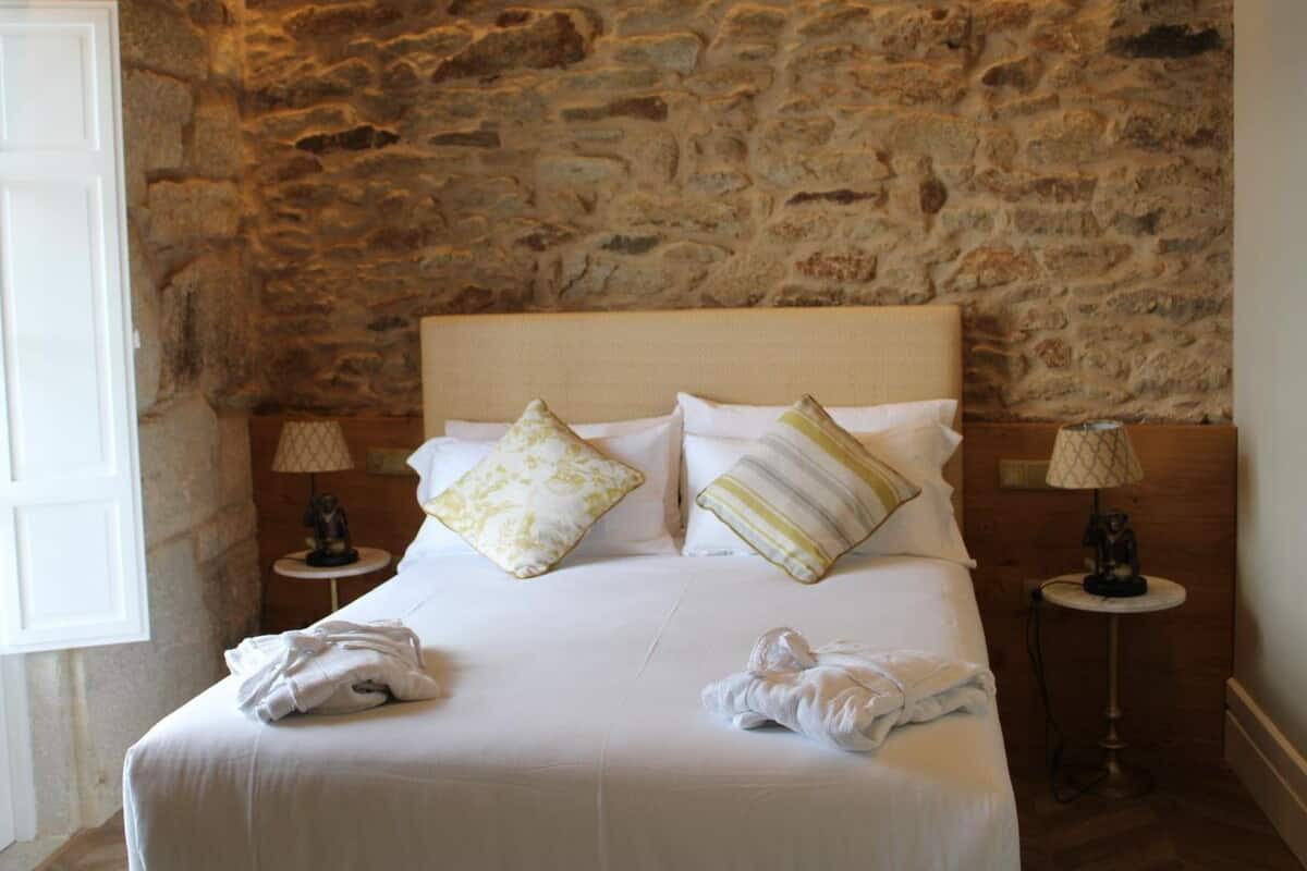 Una cama con sábanas blancas, dos almohadas y una colcha. Hay dos mesitas de noche con lámparas. La habitación tiene una pared de piedra.