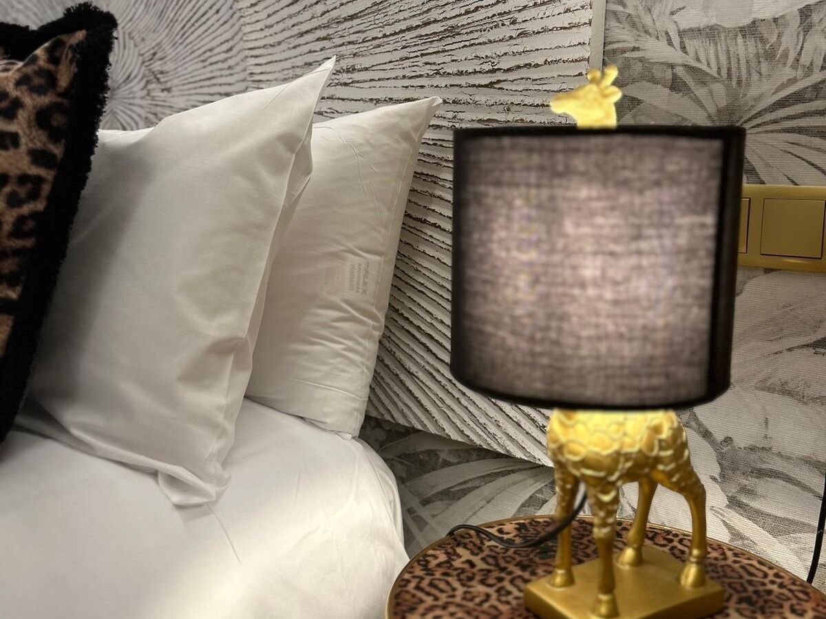 Una cama con sábanas blancas, un cojín estampado de leopardo, una lámpara con base de leopardo y una pared con diseño.