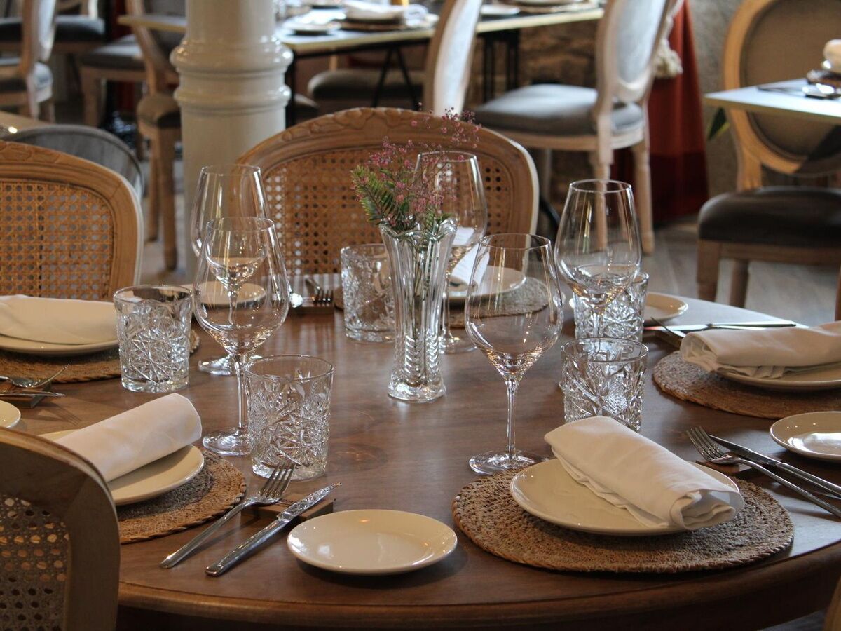 Una mesa de comedor dispuesta con varias copas de vino, platos y cubiertos, rodeada de sillas de mimbre.