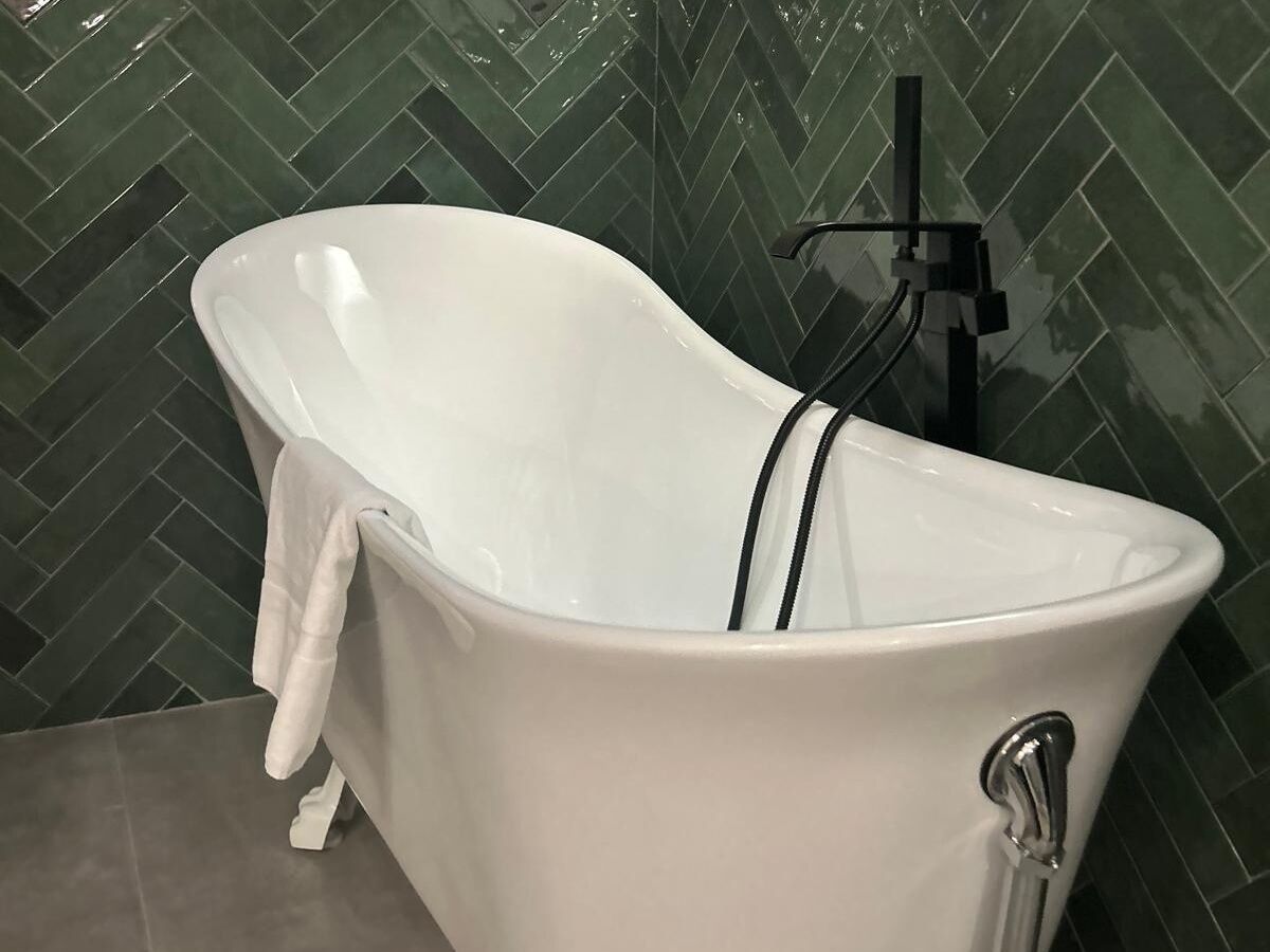 A white freestanding bathtub with black stripes, a towel, and a black faucet in a bathroom with green herringbone patterned tiles.