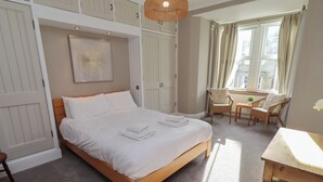 Ein ordentlich bezogenes Bett mit weißen Leinentüchern und Handtüchern, ein Korbsessel, ein kleiner Tisch und Blick auf die Stadt durch ein Fenster.