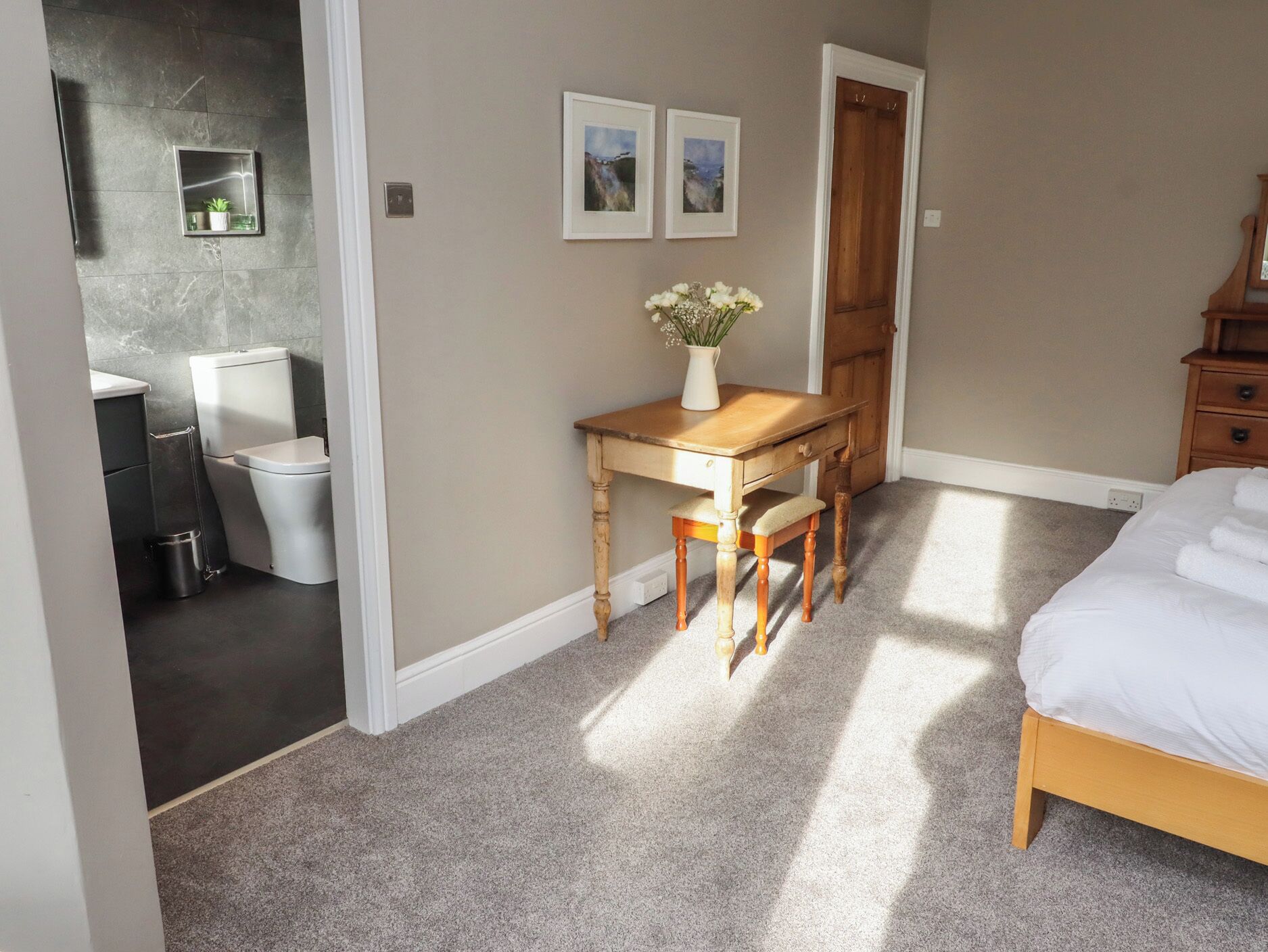 A bedroom with a bed, a wooden desk with a chair, a small table with a vase of flowers, and a door leading to a bathroom.