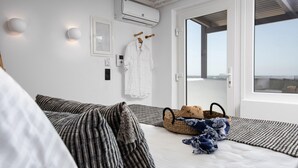 A hotel room with a bed, pillows, a bedside table, a basket, and a view of the city through a large window. - K Town Suites Naxos Penthouse Apartment (Naxos)