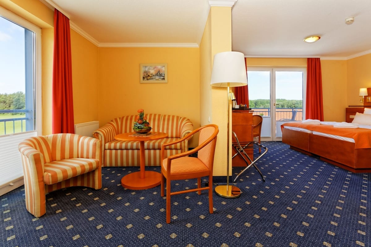Een hotelkamer met een bank, fauteuil en een kleine tafel met een lamp. Er is een bed met een witte deken en uitzicht op de omgeving door een schuifdeur.