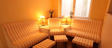A cozy living room with striped sofas and ottomans, a small table with lamps, and a window with blinds.