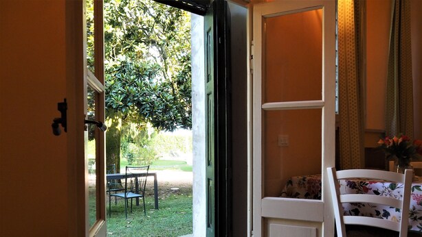 An open door leading to a patio with a table and chairs, a small garden, and a view of trees. - Magnolia Apartment (Colombare)
