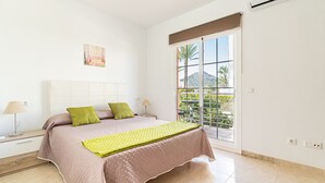 Une chambre à coucher avec un lit, deux oreillers, une table de chevet, une lampe et une fenêtre donnant sur un espace verdoyant, ainsi qu’un balcon.