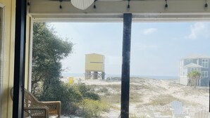 Um deck de madeira com uma cadeira de vime e cadeiras Adirondack brancas, com vista para uma praia arenosa com um buggy e um tanque de água à distância.