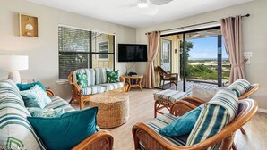 Uma sala de estar ampla com móveis de vime, uma televisão de tela plana e uma grande porta de vidro deslizante que dá para uma varanda com vista para a praia.