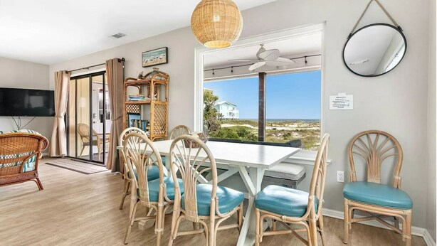 Uma sala de jantar com cadeiras de vime, uma mesa branca, uma televisão e vista para a praia.