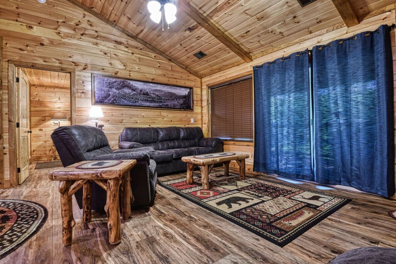 A cozy cabin interior with a wooden ceiling, a large window with blue curtains, a black leather sofa, and a rug with a geometric pattern.