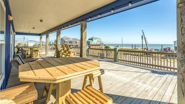 A wooden deck with a table and benches, overlooking a beach with houses and a crane in the background.