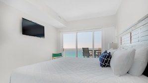 A hotel room with a large bed, a flat-screen TV, a chair, and a view of the ocean through large windows.