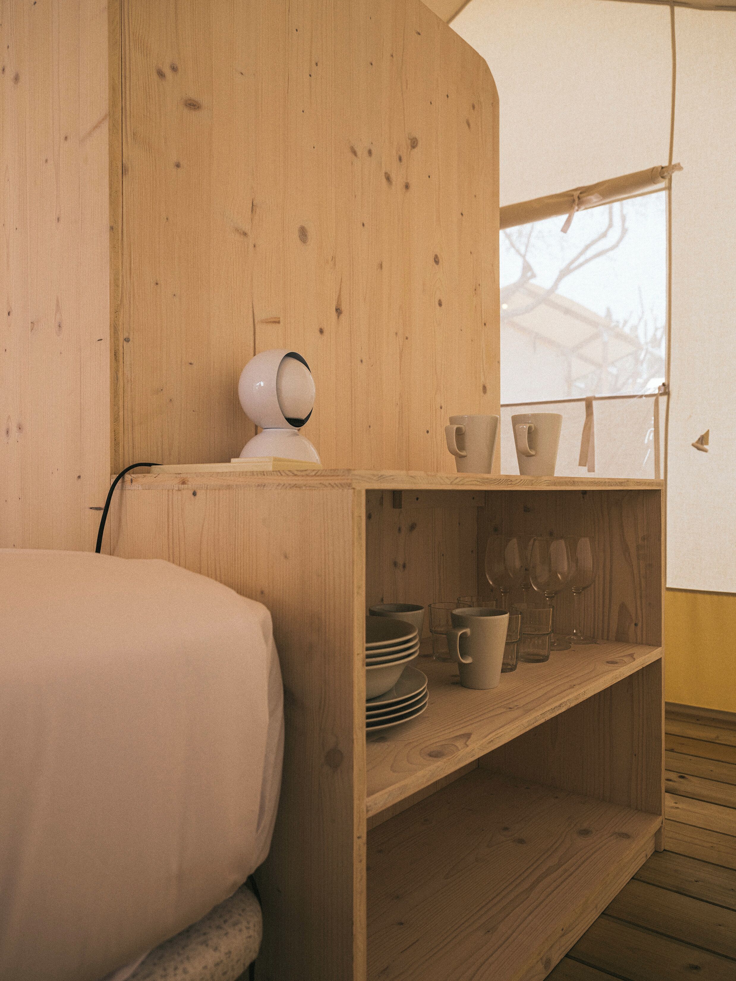 A wooden room with a bed, a shelf with cups and glasses, and a window with a view of trees.
