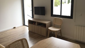 A room with a wooden floor, a TV on a wooden stand, a wicker chair, and a window with blinds.