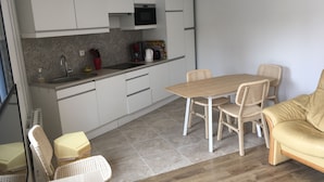 A compact kitchen with white cabinets, a microwave, and a sink. There is a dining area with a wooden table and wicker chairs. A beige sofa is placed near the dining area.