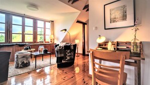 A cozy living room with wooden floors, a sofa, a coffee table, and a dining table with chairs. There are windows with curtains, a lamp, and a framed picture on the wall.