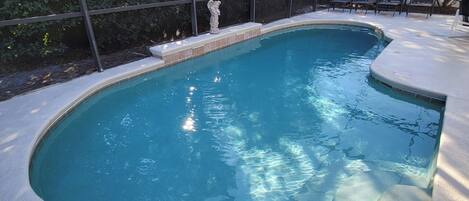 Una piscina con borde curvo, rodeada por una zona techada con sillas y una estatua.