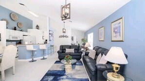 Un salón moderno con un sofá de cuero negro, una mesa de centro de vidrio y una alfombra azul. Hay una zona de cocina con armarios blancos y una mesa de comedor con taburetes de bar.