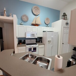Una cocina con armarios blancos, microondas, estufa, refrigerador y fregadero.