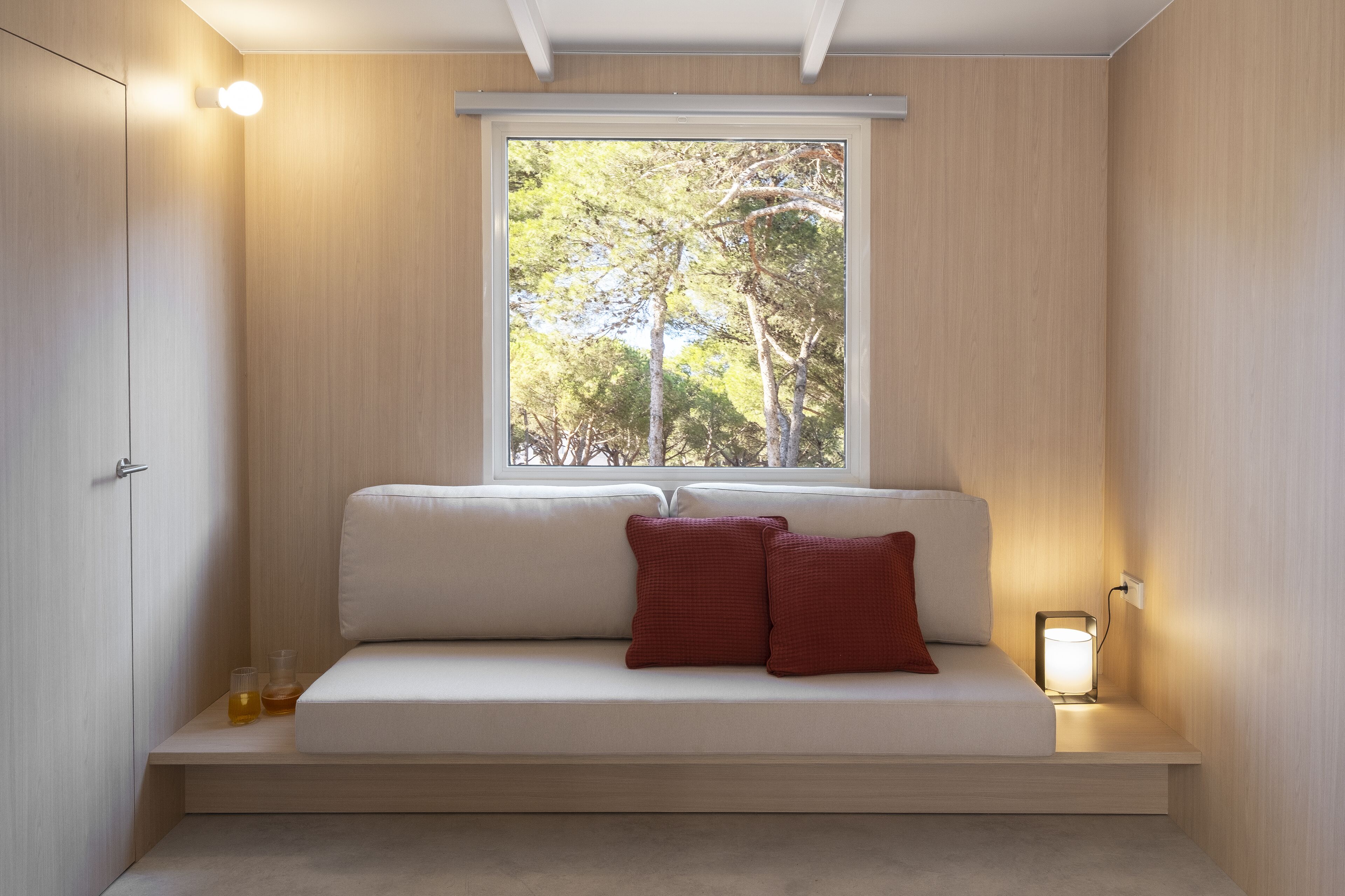 A modern room with a built-in bench, a window with a view of trees, and a bedside table with a lamp.