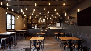 Un interior de restaurante con mesas de madera y sillas negras, una zona de bar y bombillas colgantes.