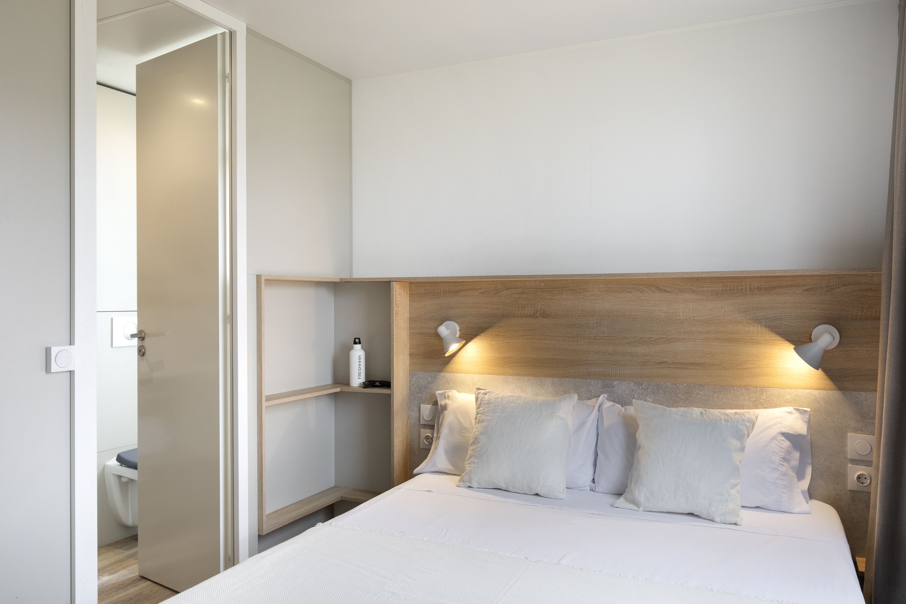 Una habitación de hotel moderna con cabecera de madera, ropa de cama blanca y una mesita de noche con una botella y una lámpara.