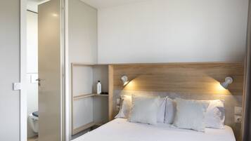 Una habitación de hotel moderna con cabecera de madera, ropa de cama blanca y una mesita de noche con una botella y una lámpara.
