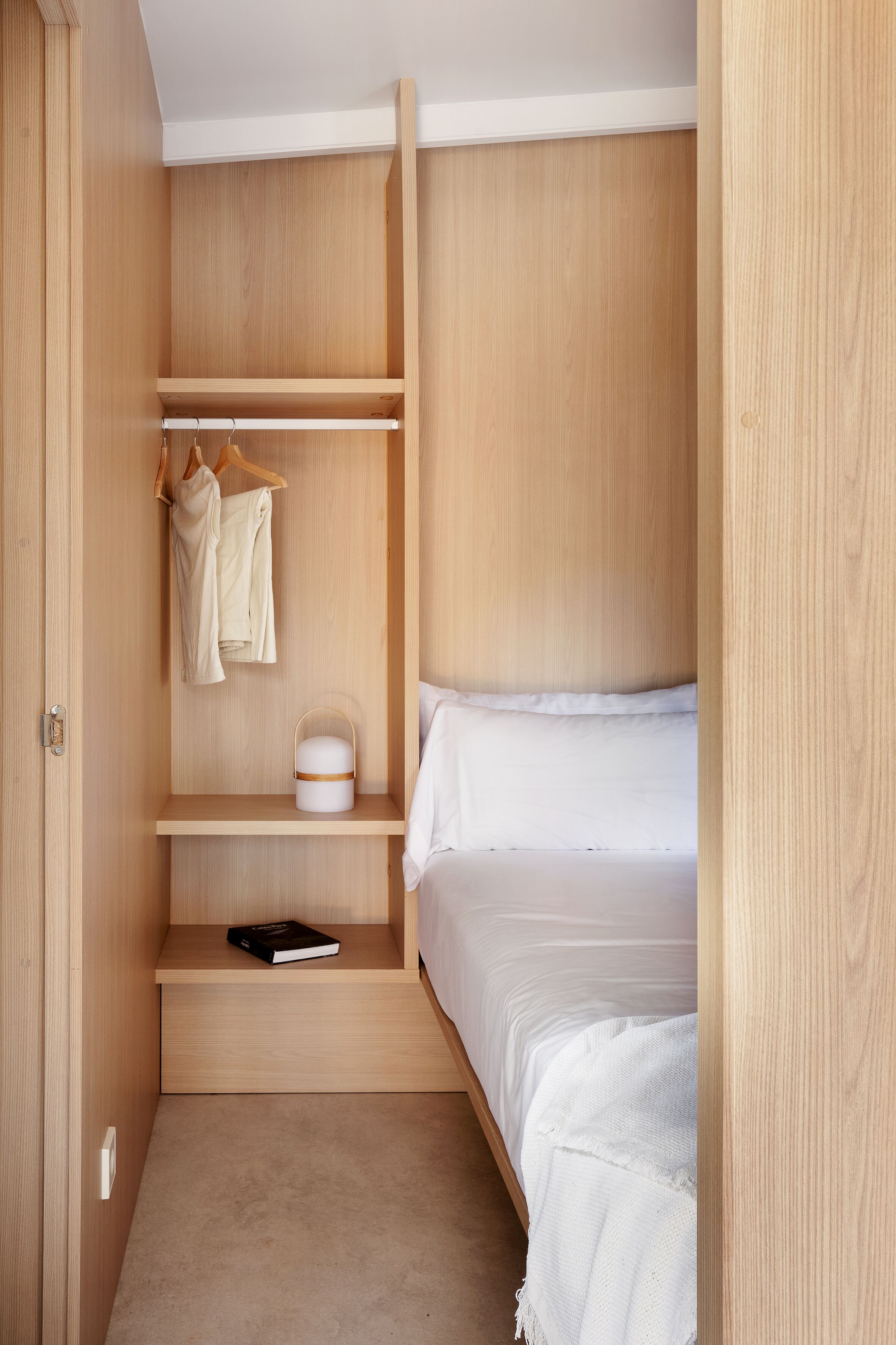 Un dormitorio acogedor con un armario de madera, una cama con ropa blanca y una mesita de noche con un libro y un recipiente blanco.