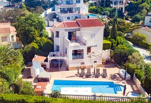 En moderne villa med swimmingpool, omgivet af frodig vegetation og andre boliger.