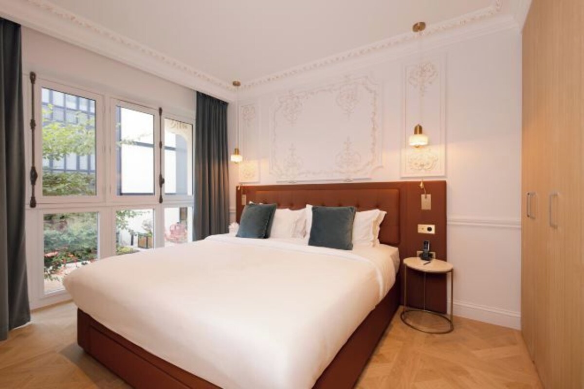 Una cama grande con ropa de cama blanca y almohadas oscuras, una mesita de noche pequeña con una lámpara, y una ventana con cortinas.