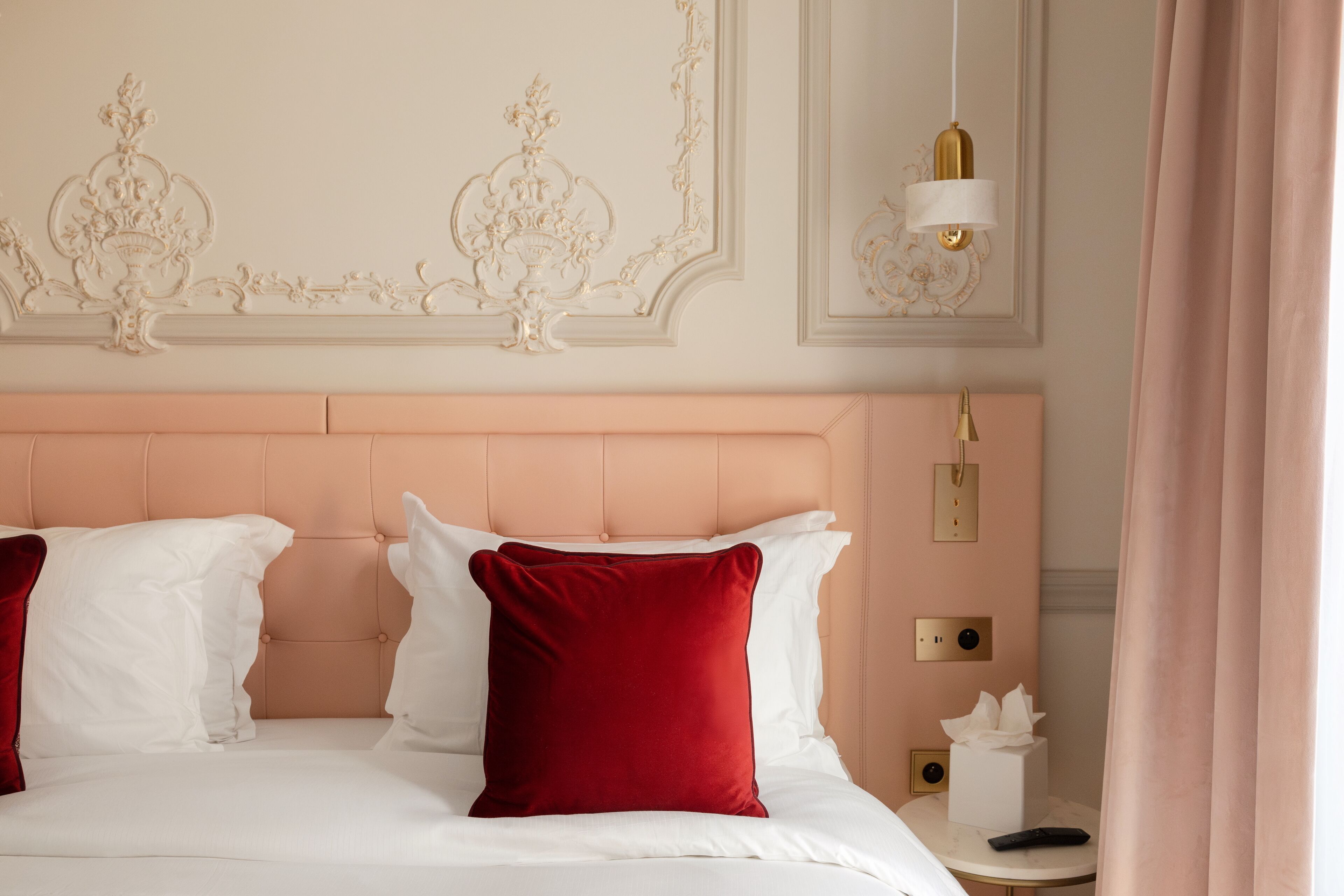 Camera d'albergo con testiera imbottita, un letto bianco con cuscini rossi e bianchi, un comodino con un telecomando e un lampadario a parete.