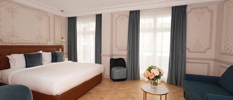 A hotel room with a large bed, a small round table with a vase of flowers, a blue armchair, and a grey armchair.