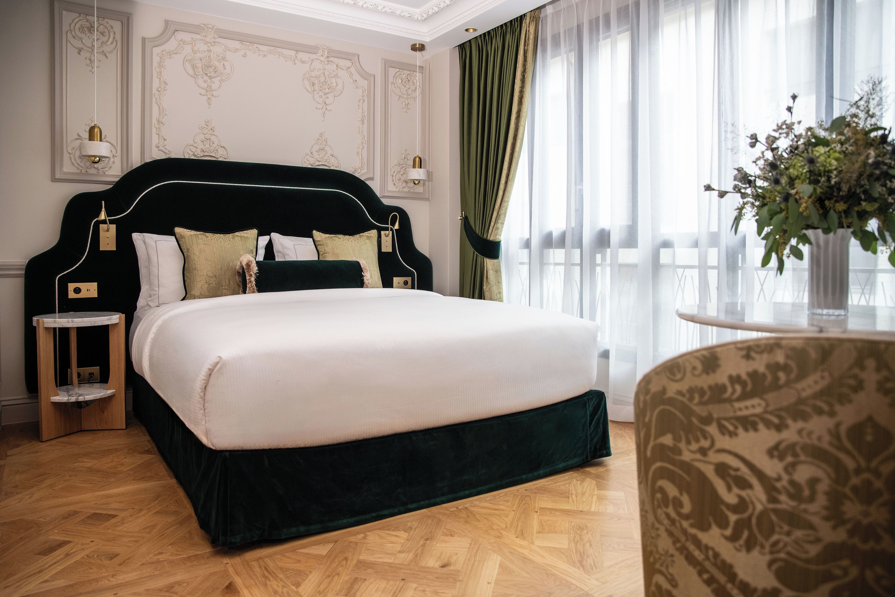 Una cama con cabecera oscura, ropa de cama blanca y varias almohadas. Hay una mesita de noche con una lámpara y una pequeña mesa redonda con un florero. La habitación tiene suelo de madera y un ventanal con cortinas traslúcidas.