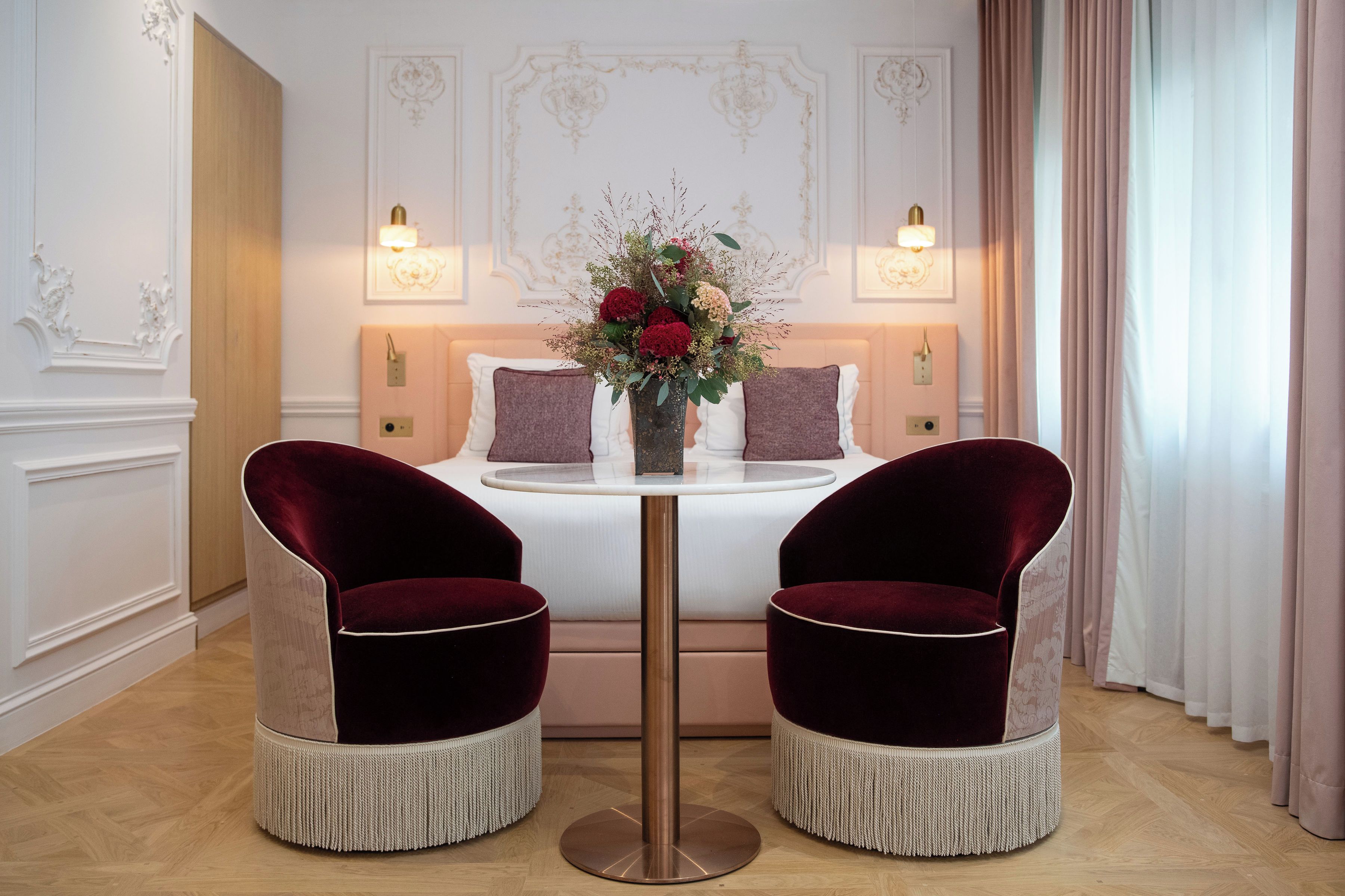 A hotel room with a bed, two armchairs, a round table with a vase of flowers, and a view of the room's decor.