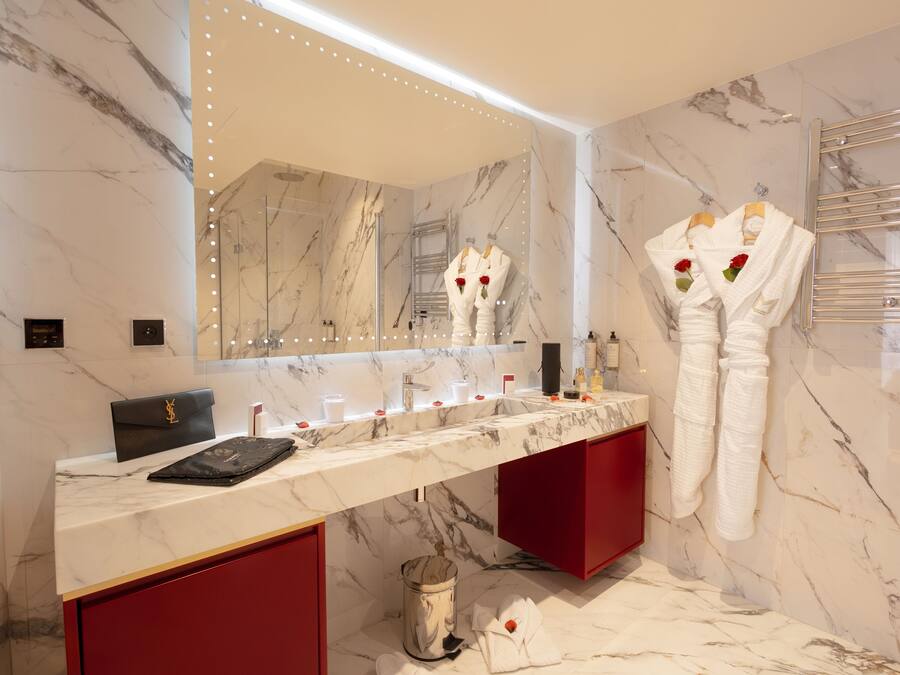 A modern bathroom with a marble vanity, a large mirror, and white robes with roses on the towel rack.