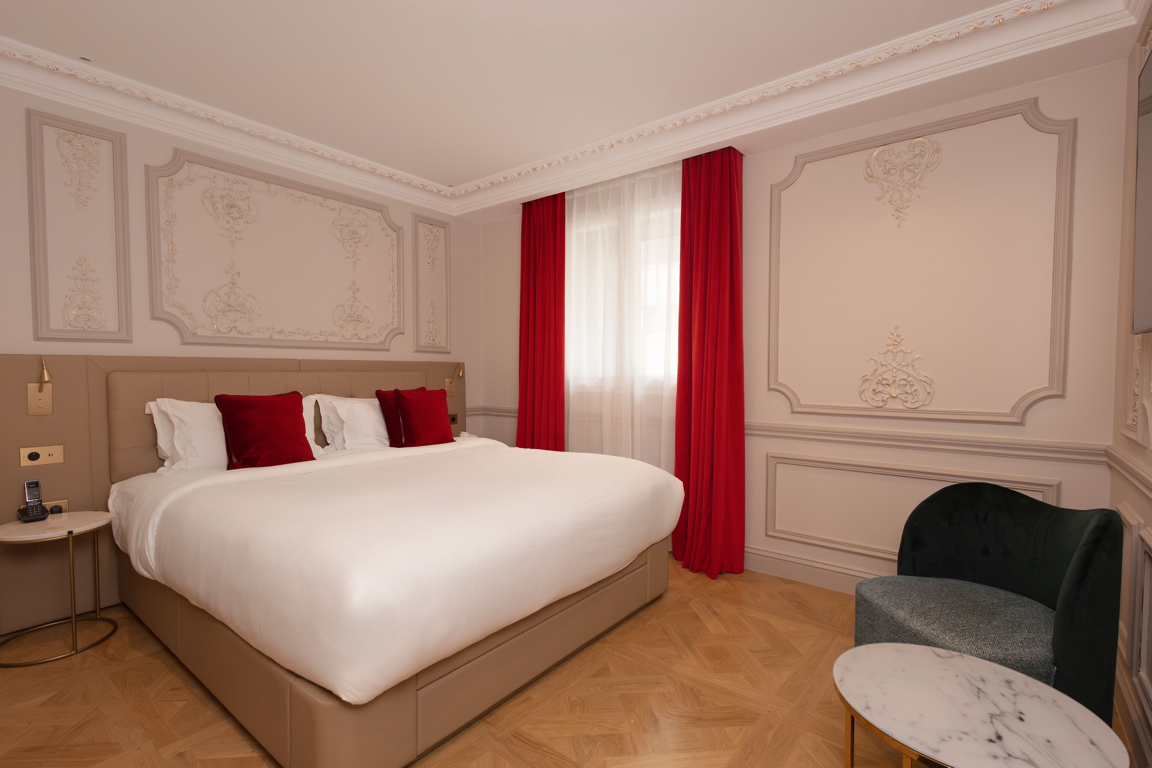 A hotel room with a large bed, red curtains, a small table, and a chair.