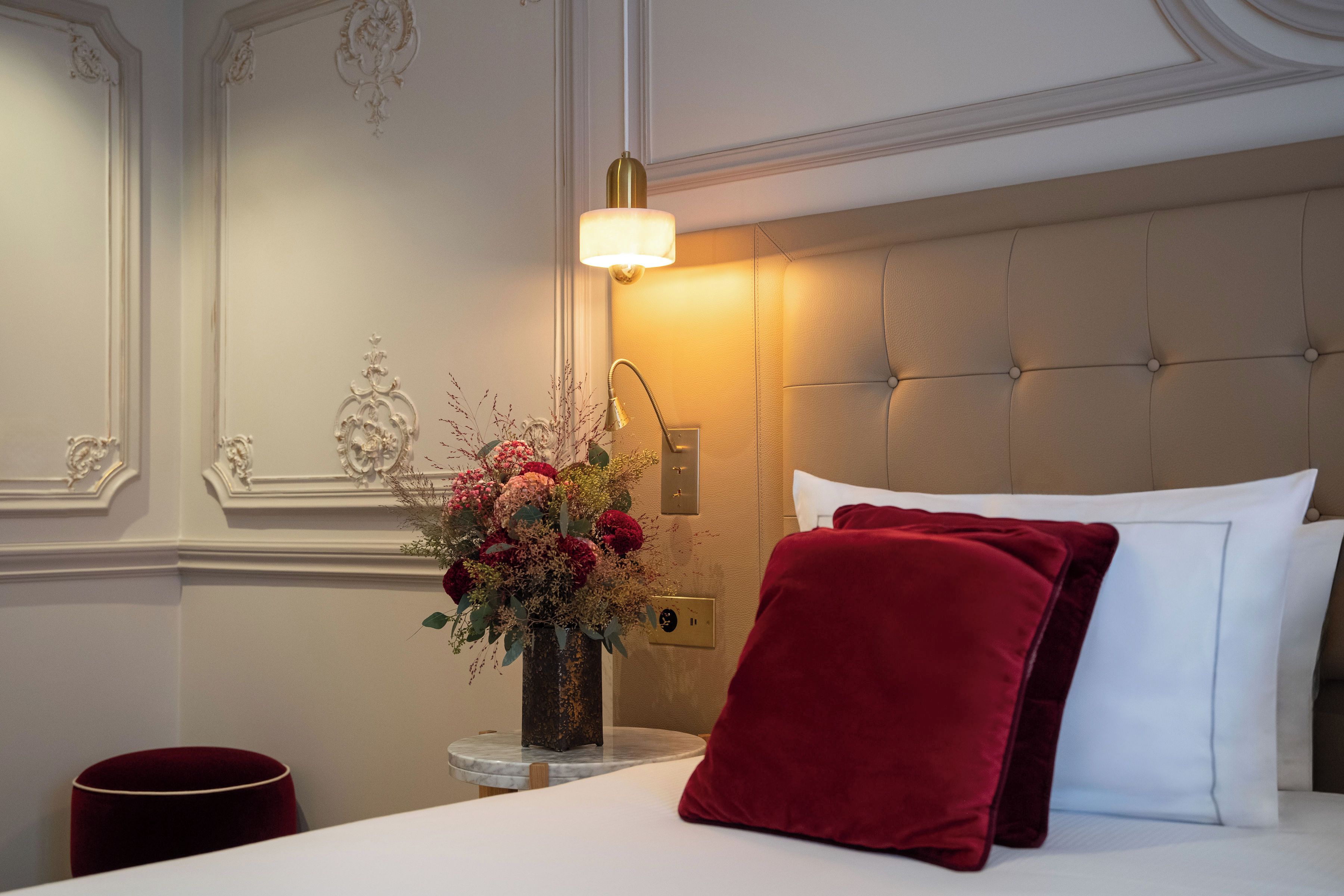 A hotel room with a bed, a vase of flowers, a lamp, and a red pillow.