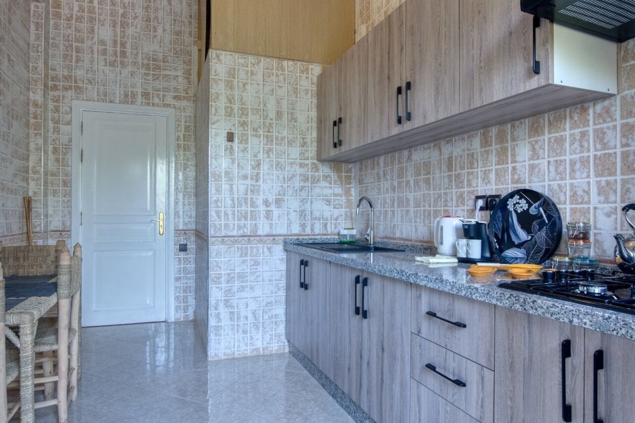 A kitchen with a tiled backsplash, wooden cabinets, a countertop with various appliances, and a dining area with a chair and table.