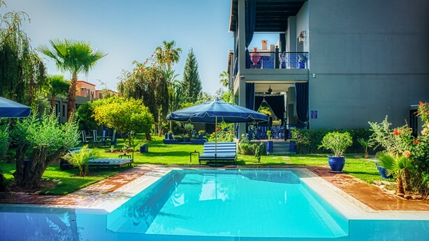 Una piscina con sillas de descanso y sombrillas, rodeada de vegetación y un edificio moderno con balcón.