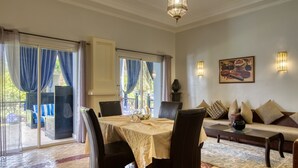 Un área de comedor con una mesa dispuesta para cuatro, un sofá con cojines, y vista a través de un ventanal a un espacio exterior con asientos.