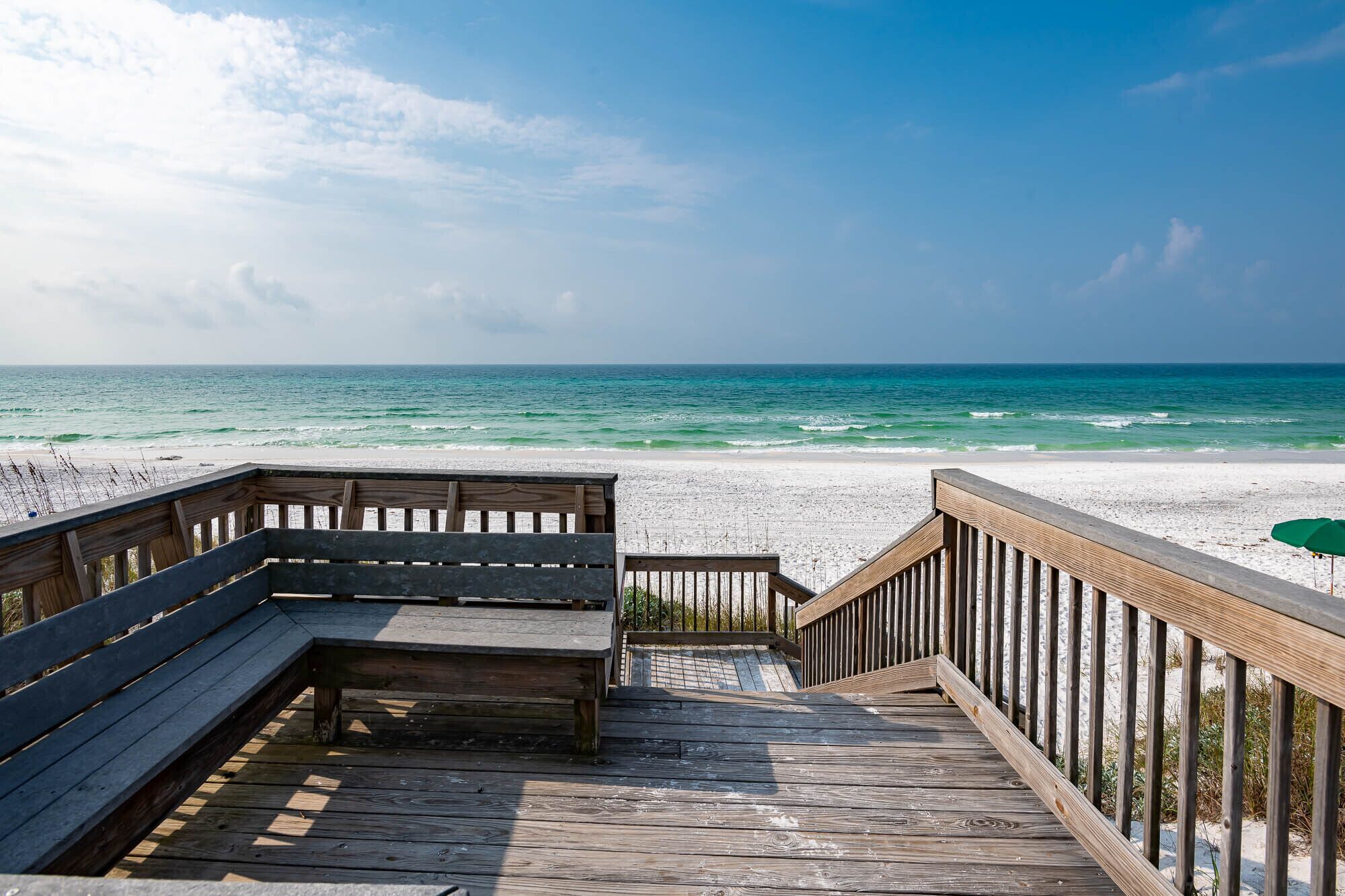 A wooden deck leading to a beach with a bench and a green umbrella.