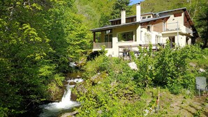 A house with a waterfall in the foreground, surrounded by lush greenery and a mountainous backdrop. - The River Mountain House at 1,000m altitude in the Pyrenées-Orièntales (Nohèdes)