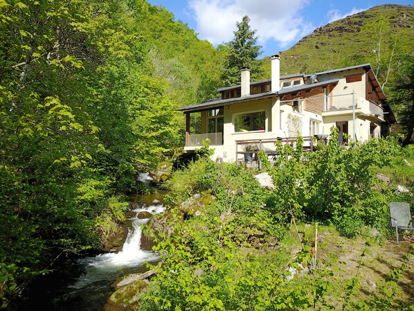 A house with a waterfall in the foreground, surrounded by lush greenery and a mountainous backdrop. - The River Mountain House at 1,000m altitude in the Pyrenées-Orièntales (Nohèdes)