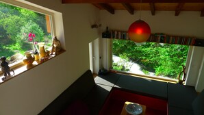 A cozy room with a large window overlooking a lush garden, a bookshelf filled with books, and a wooden ceiling. - The River Mountain House at 1,000m altitude in the Pyrenées-Orièntales (Nohèdes)