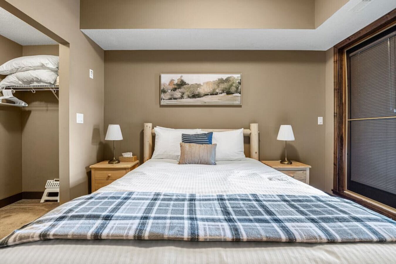 A bedroom with a bed, two bedside tables, a nightstand, a window with blinds, and a painting on the wall.