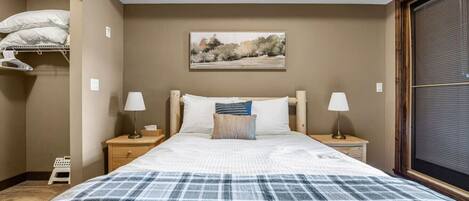 A bedroom with a bed, two bedside tables, a nightstand, a window with blinds, and a painting on the wall.